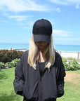 Person wearing a black jacket and black baseball cap standing on grass with ocean in the background