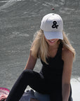 Woman sitting on a tennis court wearing a white cap with a black logo.