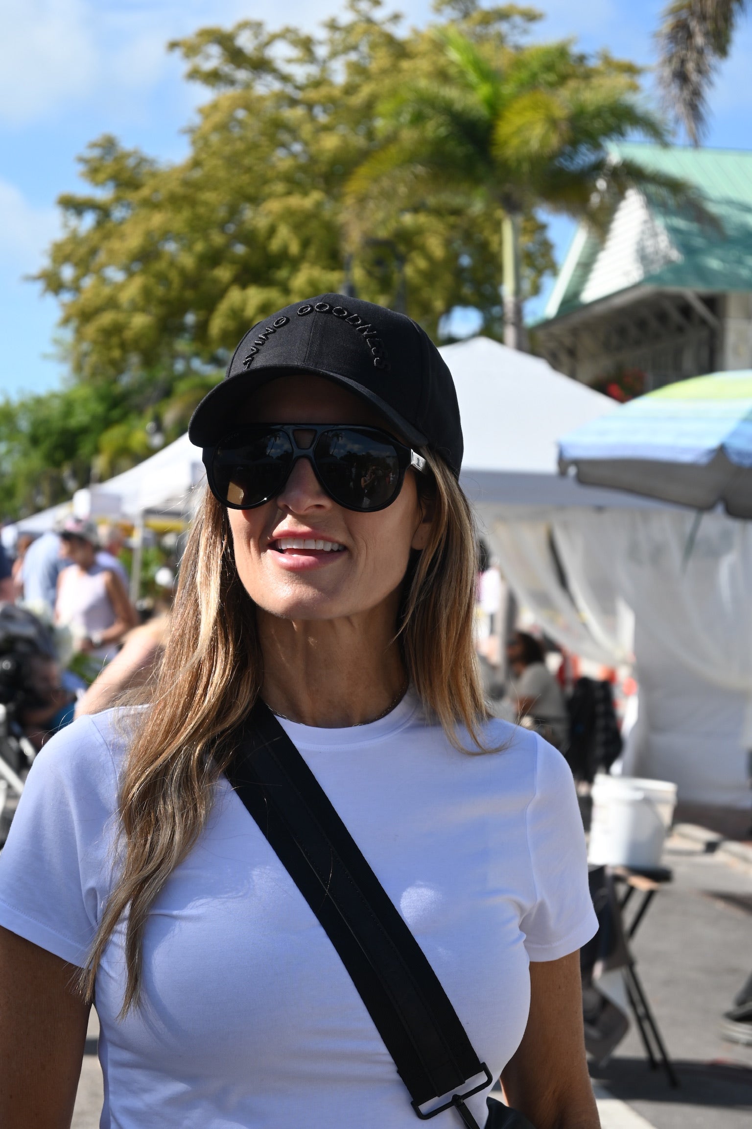 Woman wearing sunglasses and a black cap in an outdoor setting with tents and people in the background.