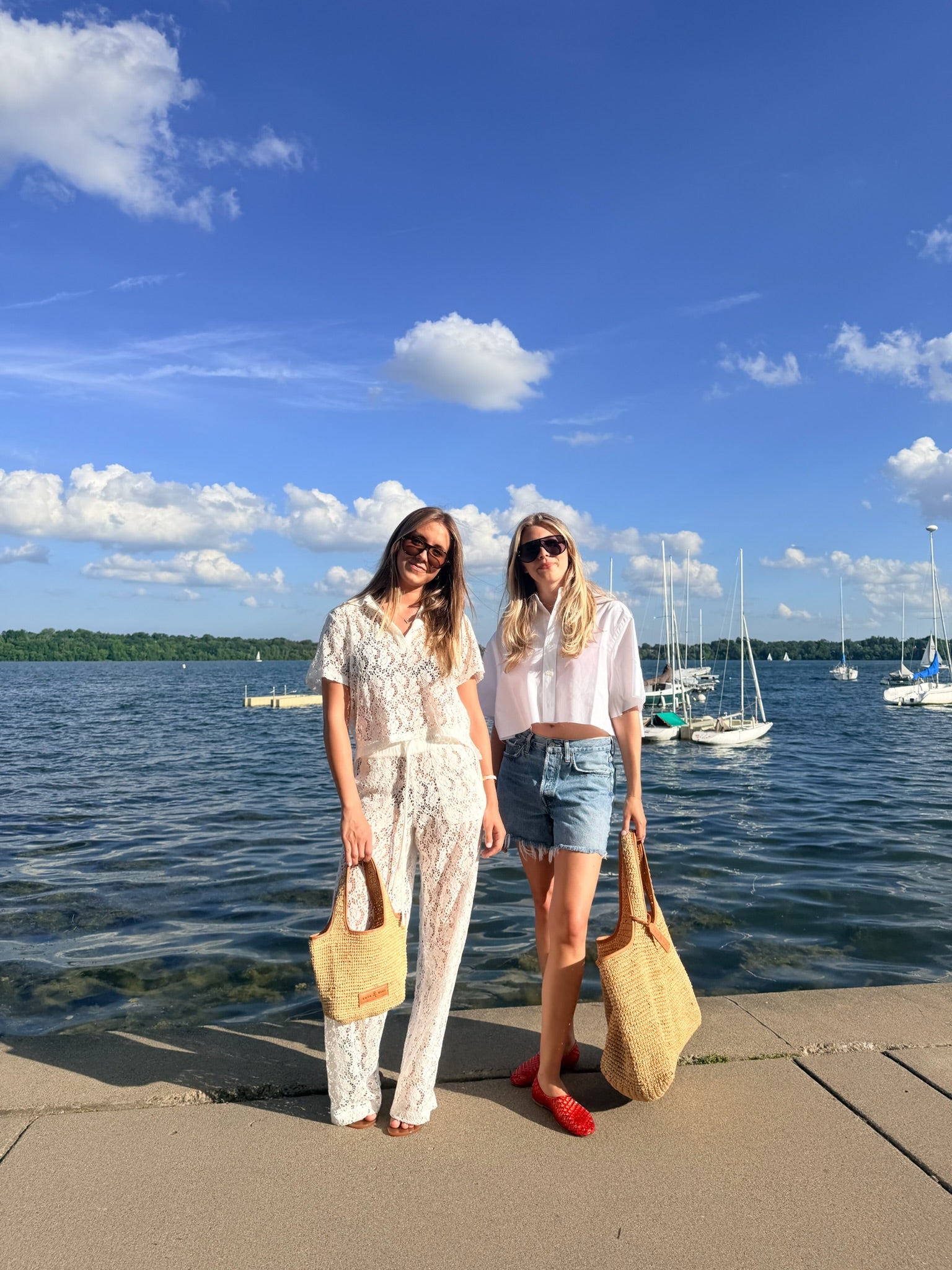 Two women standing on a concrete pier by a lake with boats and a blue sky in the background. The woman on the left wears a white lace jumpsuit and holds a beige tote bag.