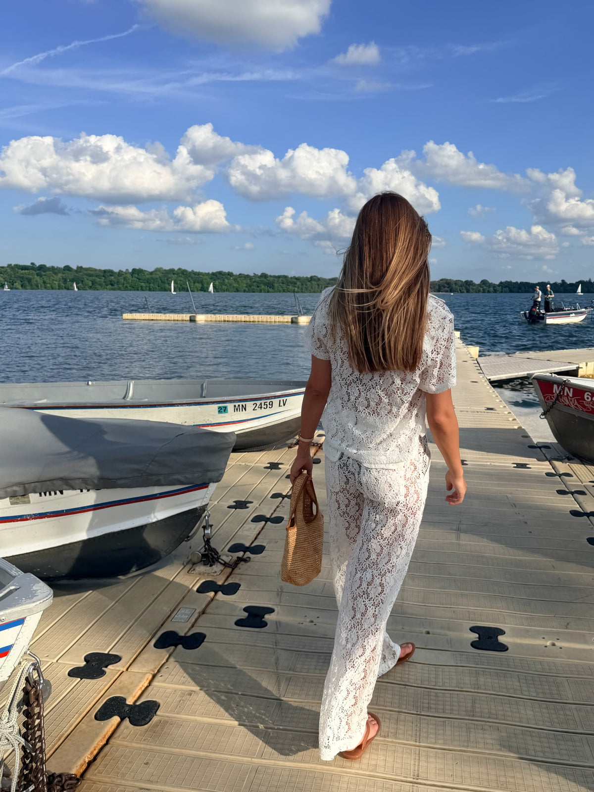 A woman viewed from behind walking on a wooden dock by the water, wearing a white lace short-sleeve top and matching white lace pants, with boats and a blue sky in the background.