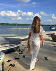 A woman viewed from behind walking on a wooden dock by the water, wearing a white lace short-sleeve top and matching white lace pants, with boats and a blue sky in the background.