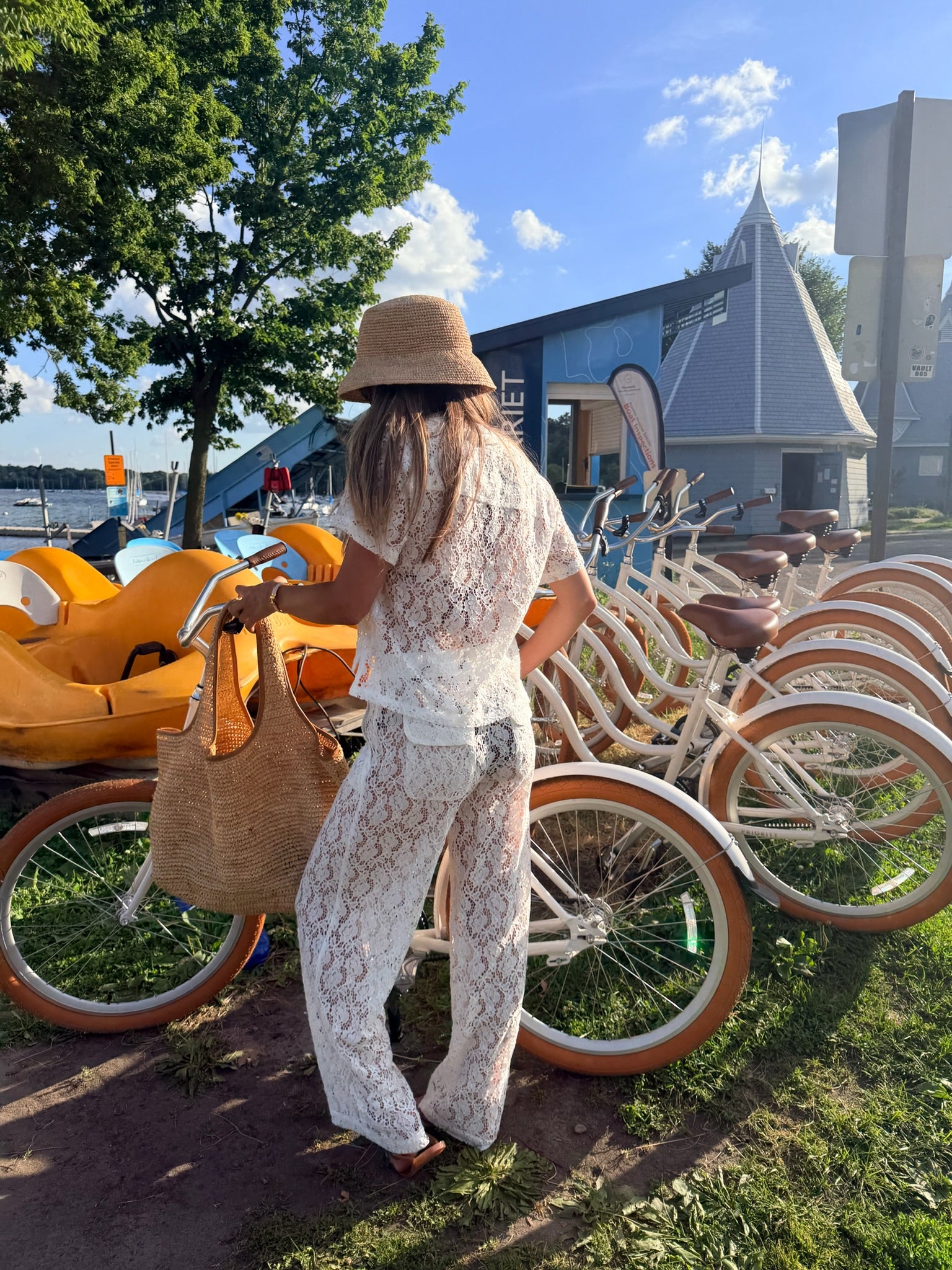 A woman wearing a matching white lace two-piece outfit and a straw hat stands outdoors near a row of cream-colored bicycles, holding a large woven tote bag.