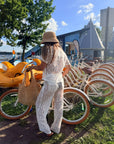 A woman wearing a matching white lace two-piece outfit and a straw hat stands outdoors near a row of cream-colored bicycles, holding a large woven tote bag.
