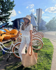 A woman wearing a white lace dress, beige straw hat, and sunglasses stands outdoors next to a white bicycle, holding a large woven straw tote bag. The background features a park setting with grass, other bicycles, and a blue sky.