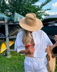A woman outdoors wearing a wide-brim straw hat, a white short-sleeve button-up shirt, and white shorts, with a red bikini top visible. She is holding a woven straw tote bag, with kayaks and trees in the background.