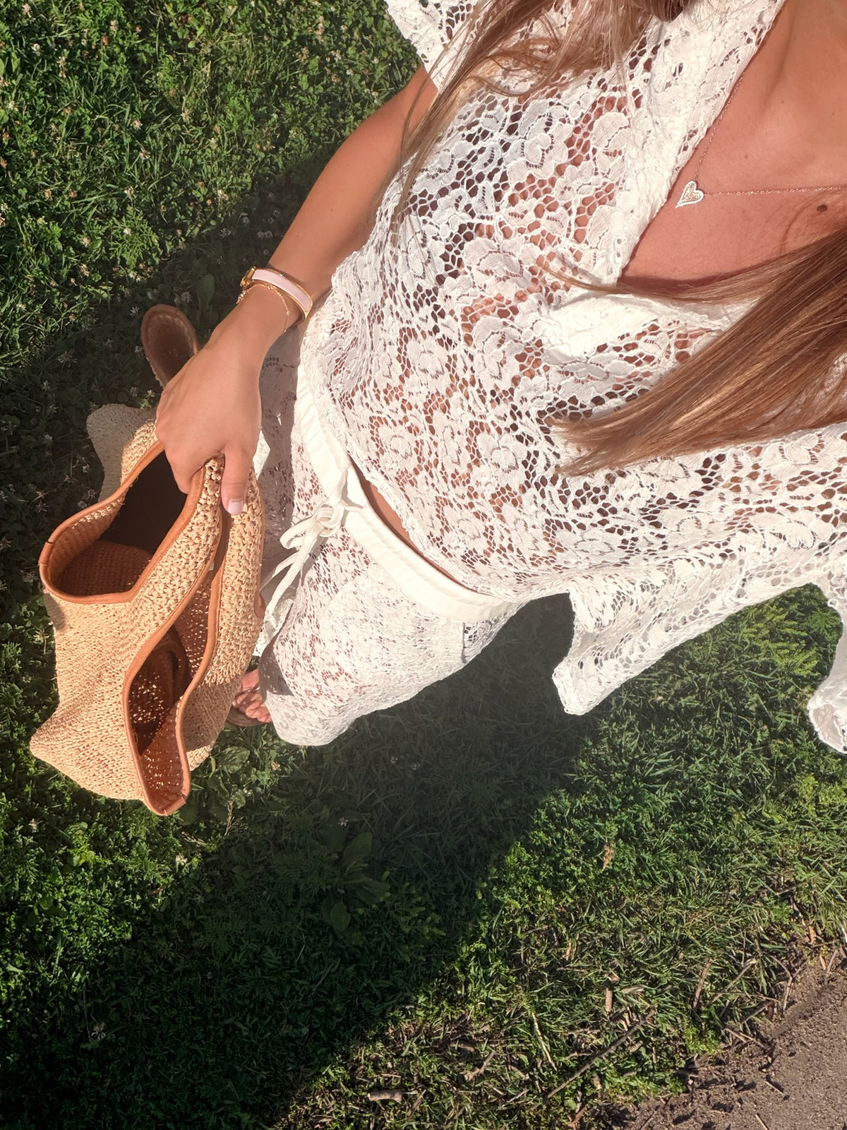 A woman outdoors on grass wearing a white lace cover-up dress and holding a woven straw bag.