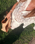 A woman outdoors on grass wearing a white lace cover-up dress and holding a woven straw bag.