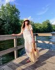 A woman stands on a wooden dock by a lake wearing a white lace maxi dress, a straw hat, and carrying a large woven straw tote bag.