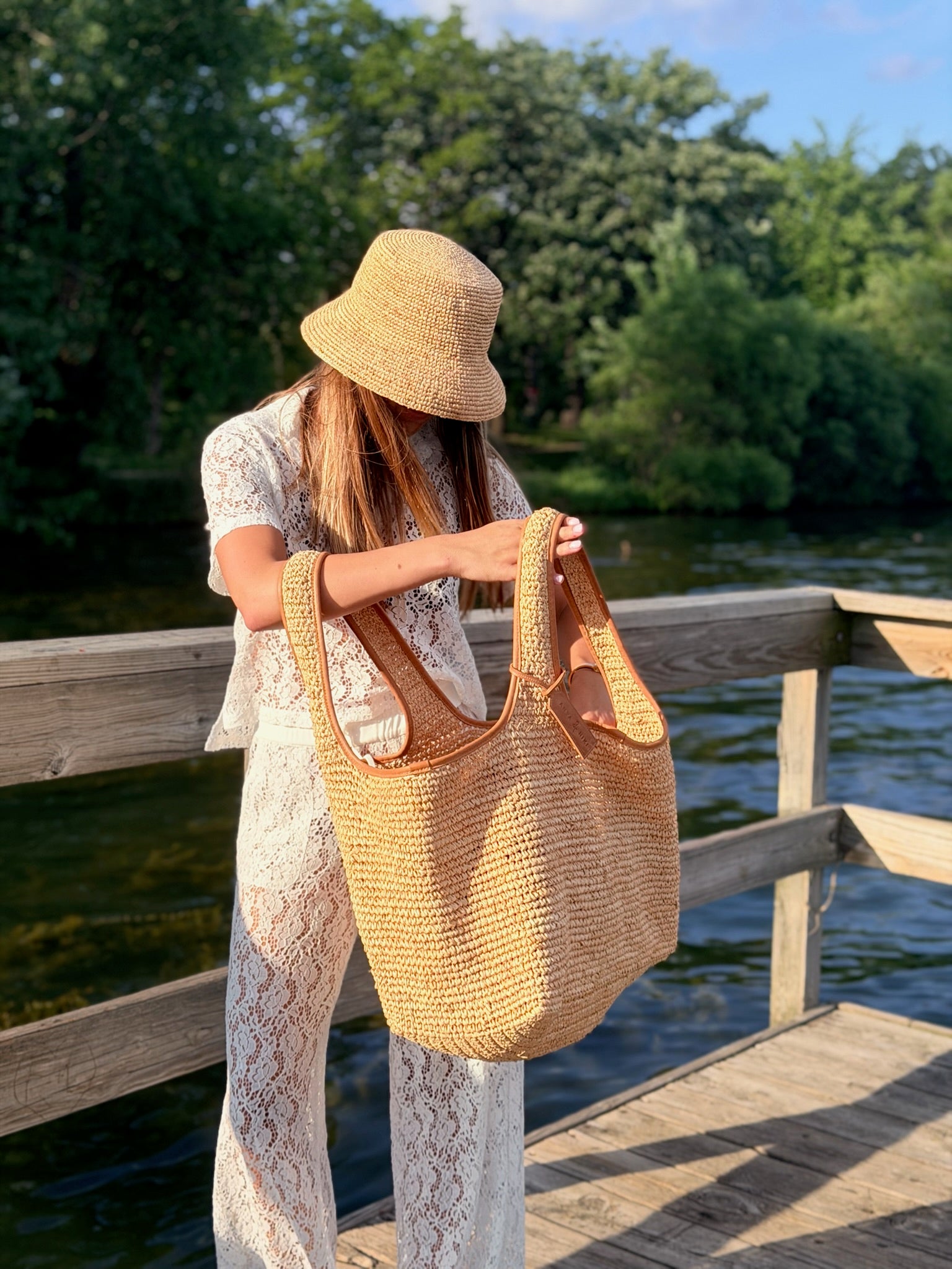 A woman wearing a straw bucket hat and a white lace outfit holds a large woven straw tote bag with brown leather handles, standing on a wooden dock by a lake with trees in the background.