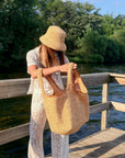 A woman wearing a straw bucket hat and a white lace outfit holds a large woven straw tote bag with brown leather handles, standing on a wooden dock by a lake with trees in the background.