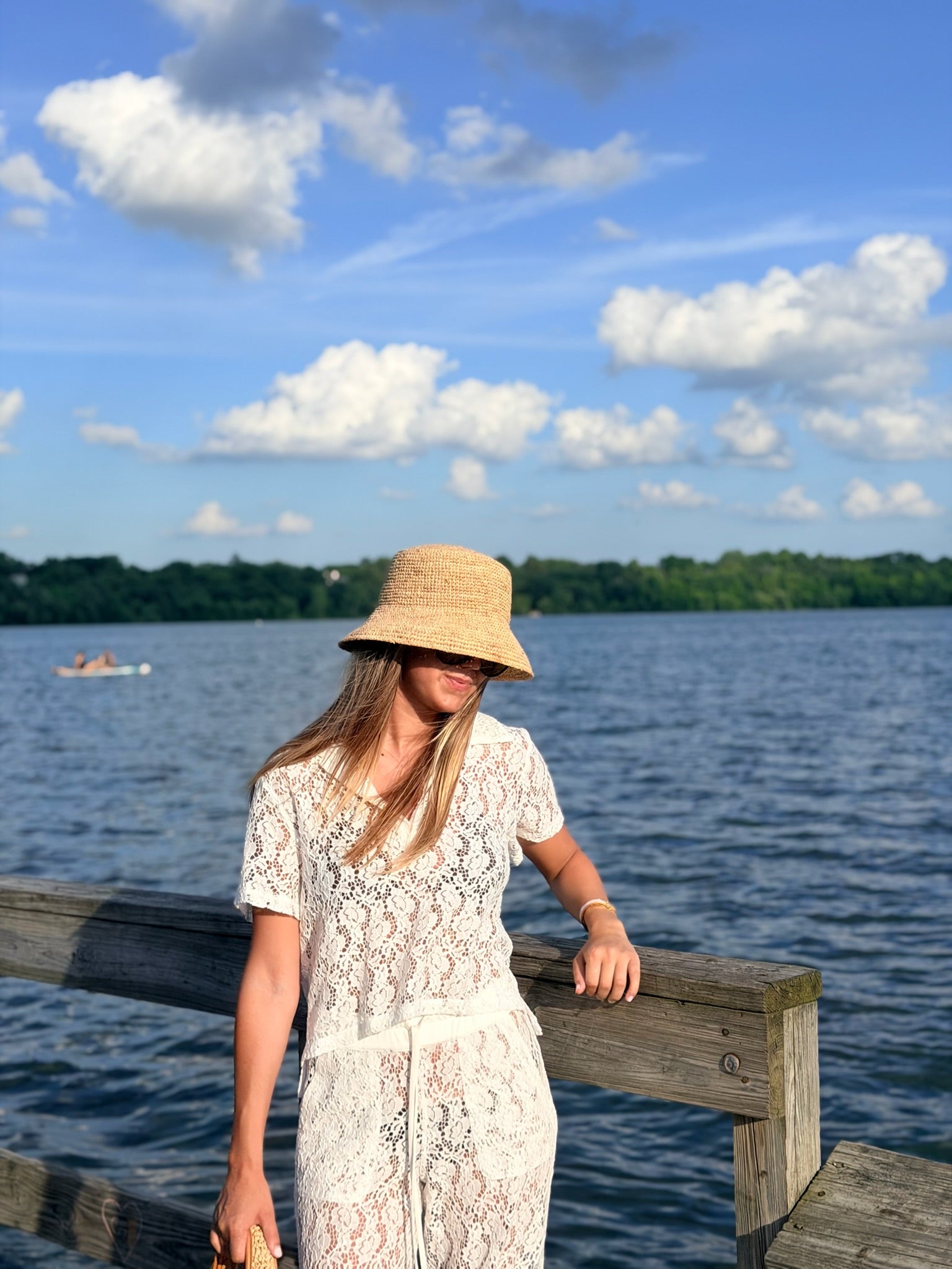 A woman wearing a straw bucket hat and a white lace short-sleeve dress stands on a wooden dock by a lake with a tree-lined shore and blue sky with clouds in the background.
