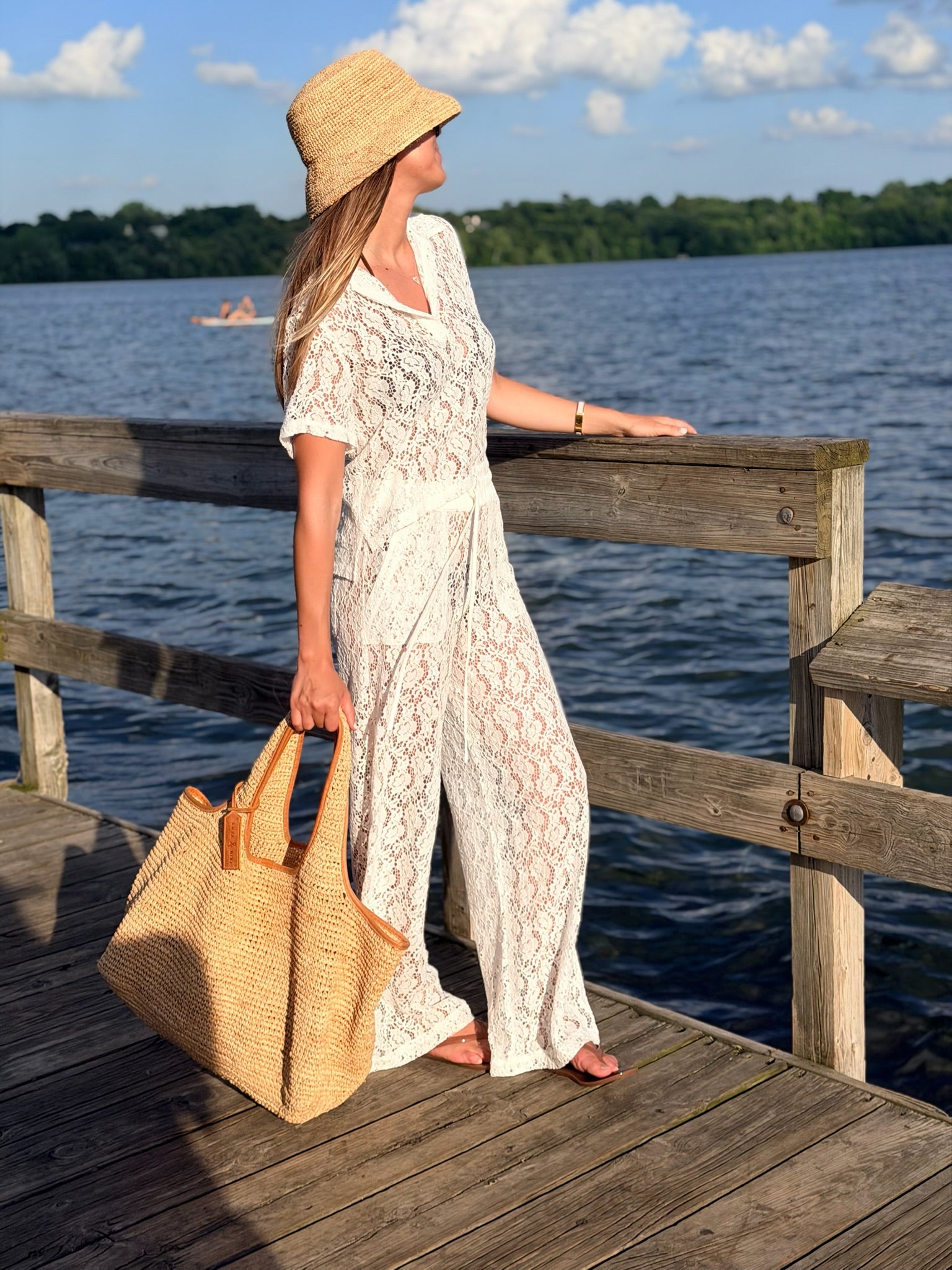 A woman stands on a wooden dock by the water wearing a white lace maxi dress, a straw hat, and carrying a large woven straw tote bag.