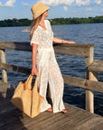 A woman stands on a wooden dock by the water wearing a white lace maxi dress, a straw hat, and carrying a large woven straw tote bag.