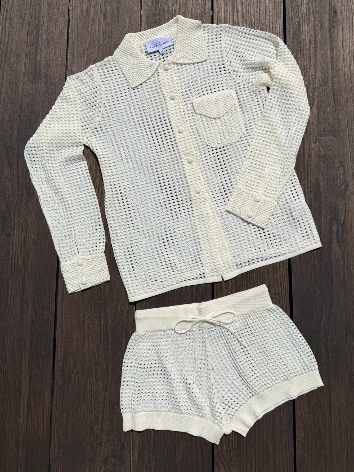 A two-piece women's knit lounge set in cream/off-white, featuring a long-sleeve button-front shirt with a collar and matching drawstring shorts, laid flat on a dark wood background.