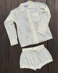 A two-piece women's knit lounge set in cream/off-white, featuring a long-sleeve button-front shirt with a collar and matching drawstring shorts, laid flat on a dark wood background.
