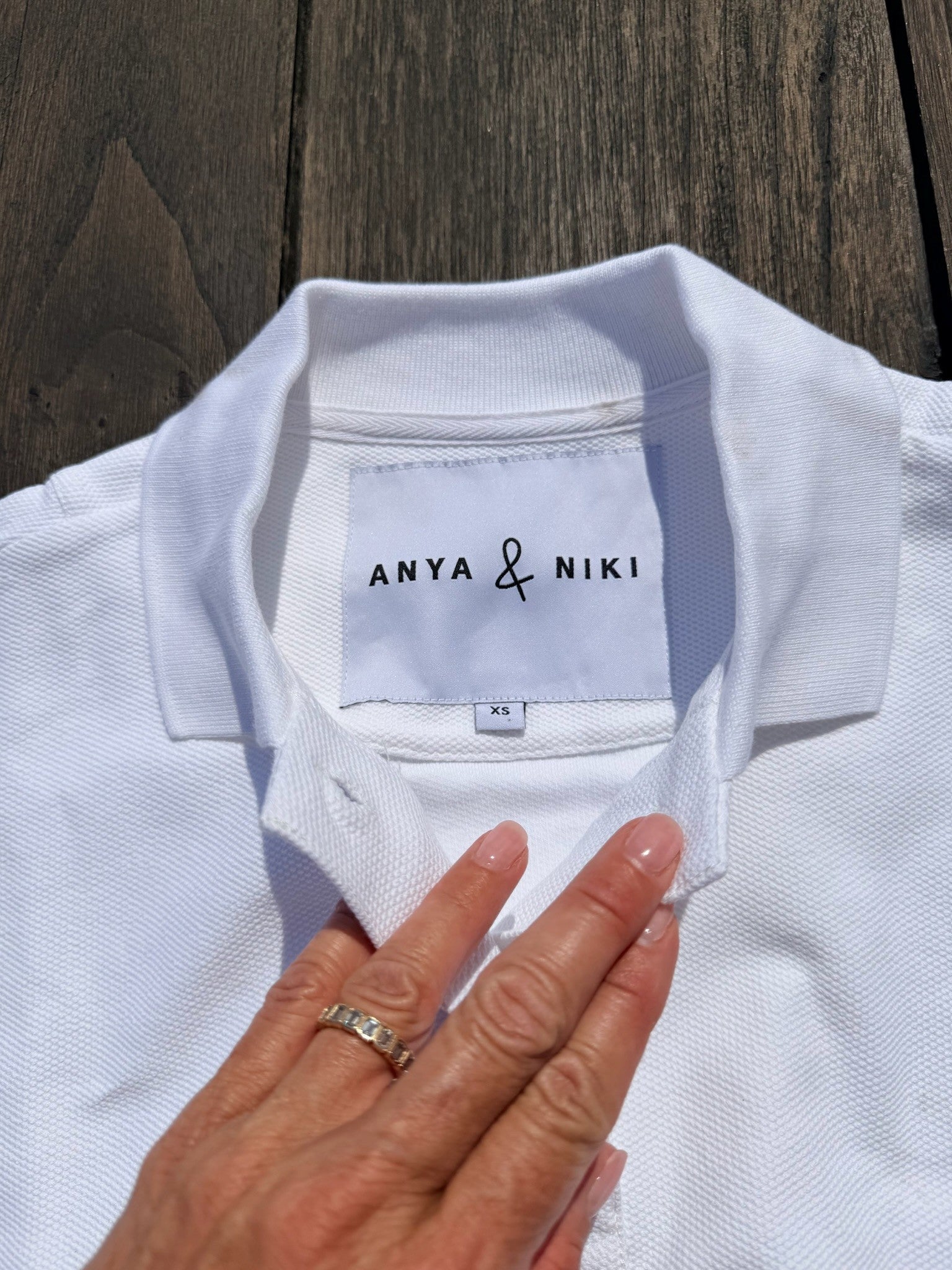 A close-up of a white polo shirt laid flat on a dark wooden surface, with a hand touching the collar area. The inside neck label reads ANYA & NIKI.
