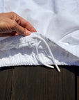 A close-up of a white textured drawstring garment, showing a hand pulling the drawstring at the elasticized waistband, set against a dark wooden background.
