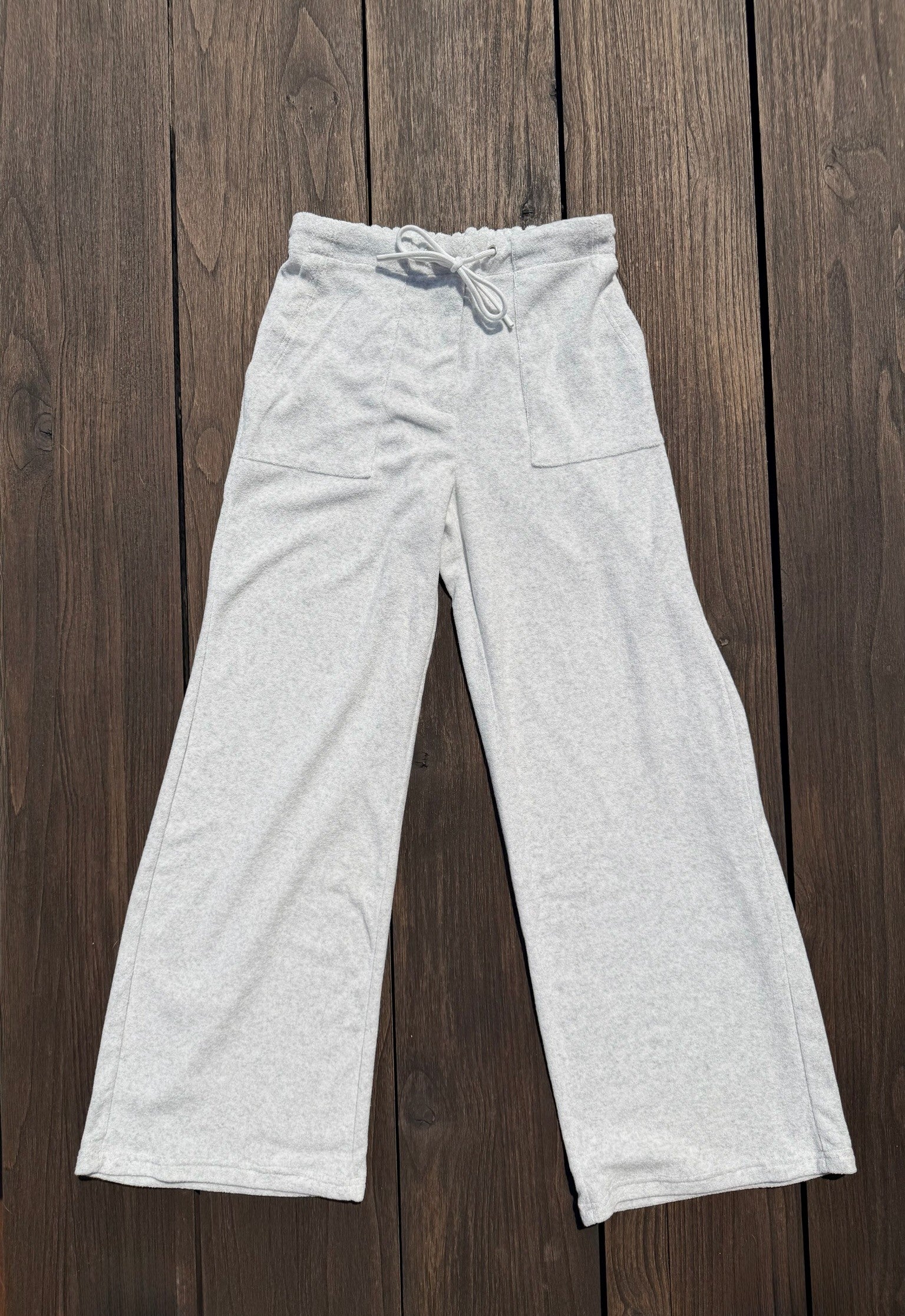 Light heather gray wide-leg sweatpants with a drawstring waistband and side pockets, laid flat on a dark brown wooden plank background.