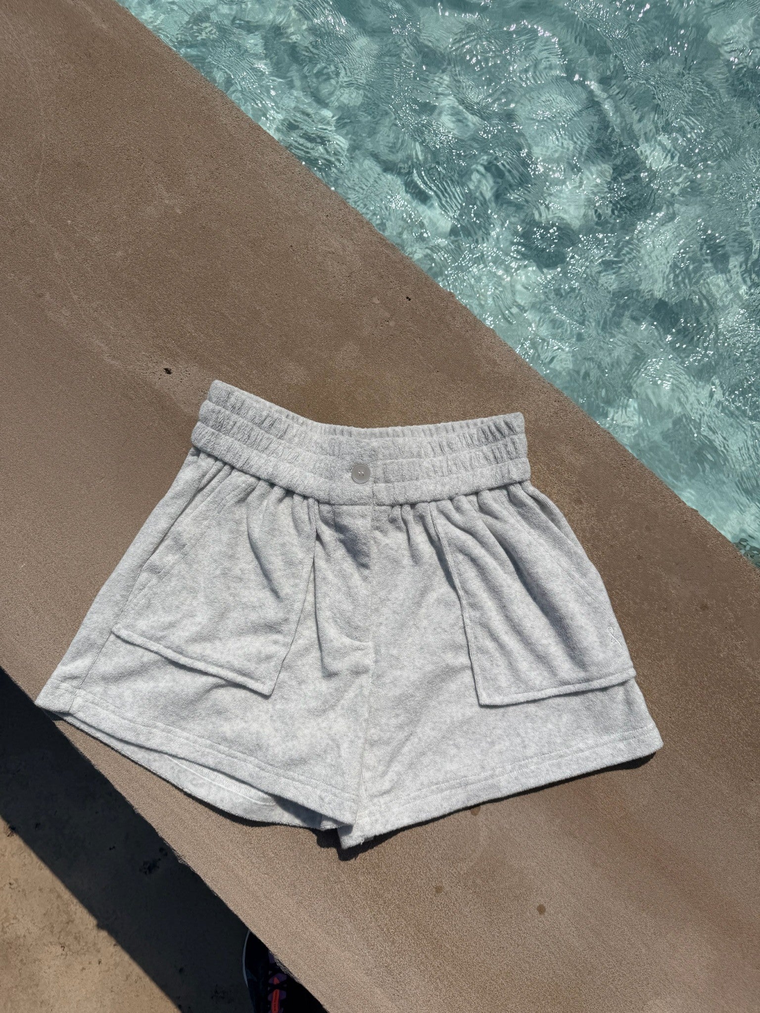 Light heather gray athletic shorts with an elastic waistband and side pockets, laid flat on a concrete surface next to a swimming pool.