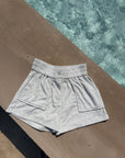 Light heather gray athletic shorts with an elastic waistband and side pockets, laid flat on a concrete surface next to a swimming pool.