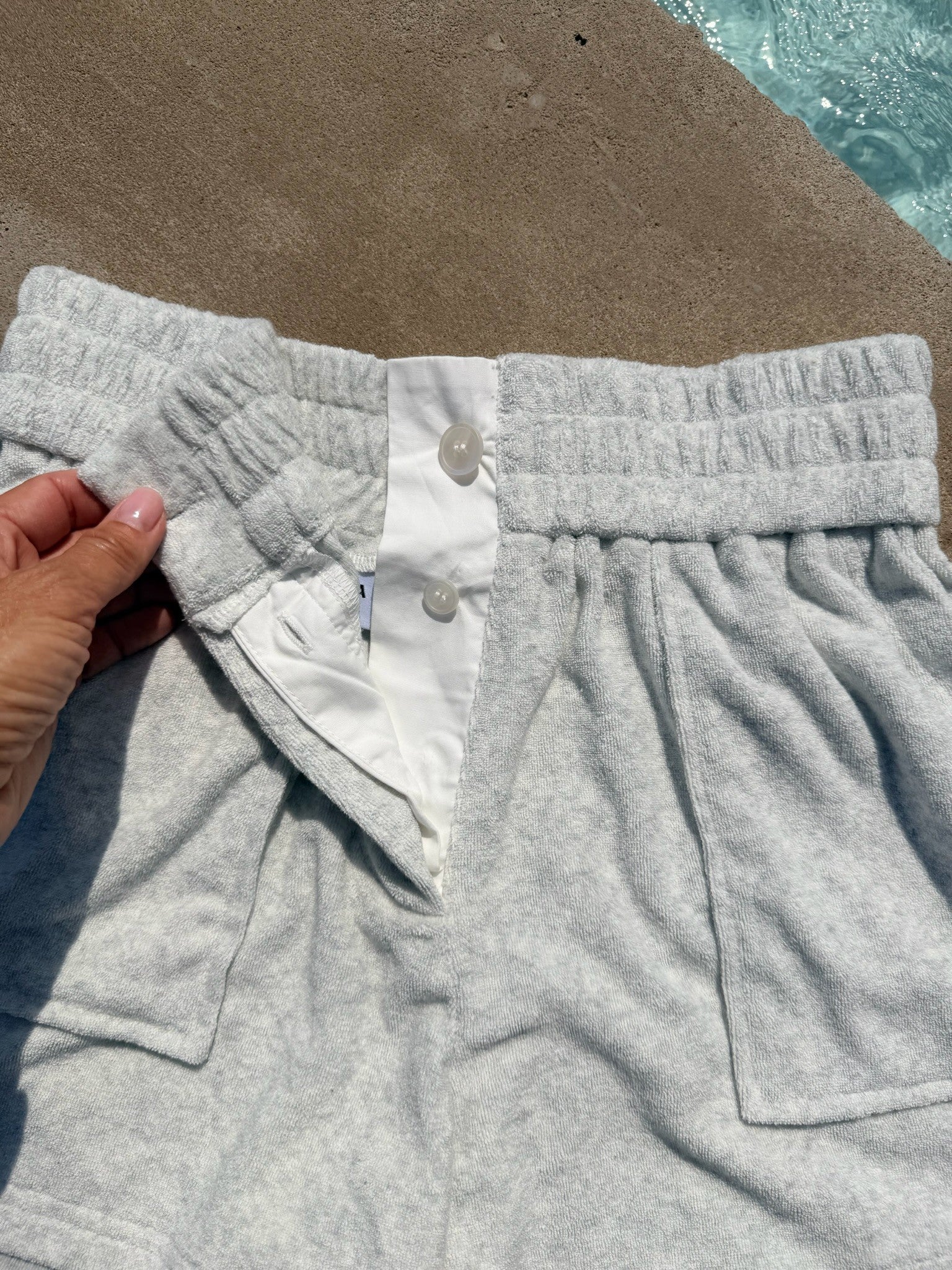 Close-up of light gray heather sweatpants with a hand holding the waistband, showing a white inner lining and snap-button closure, with a poolside concrete surface in the background.