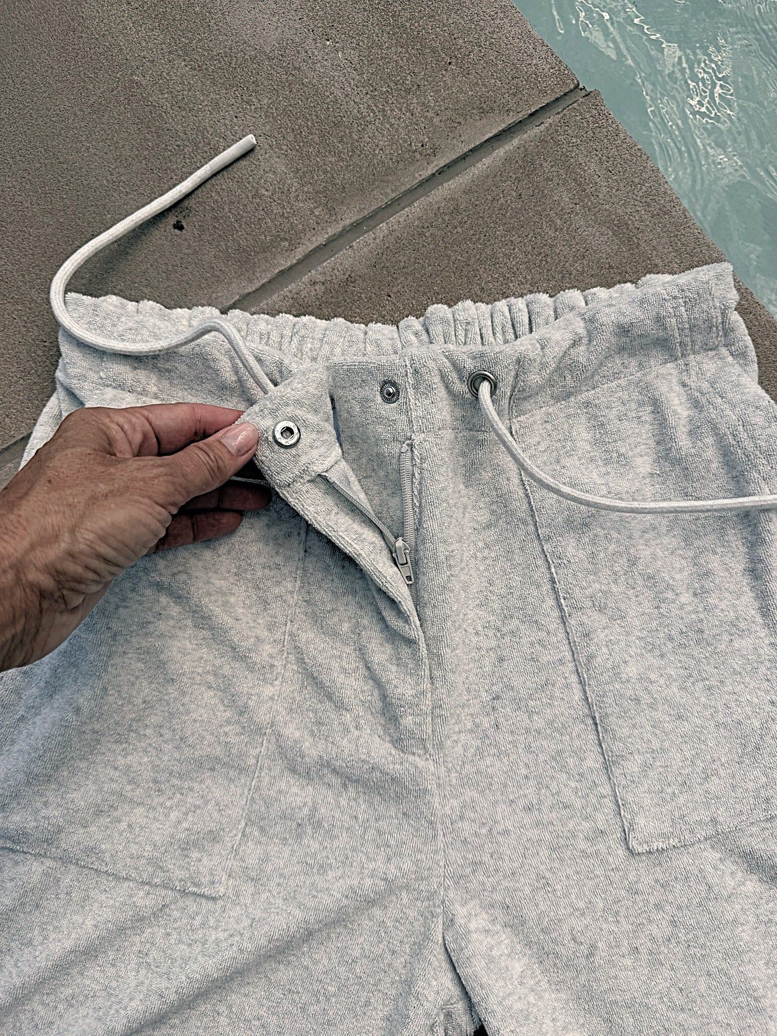 A close-up of light gray heather sweatpants with a hand adjusting the drawstring waist. The shorts are shown outdoors next to a concrete surface and water, featuring a front zipper, drawstrings, and a side pocket.