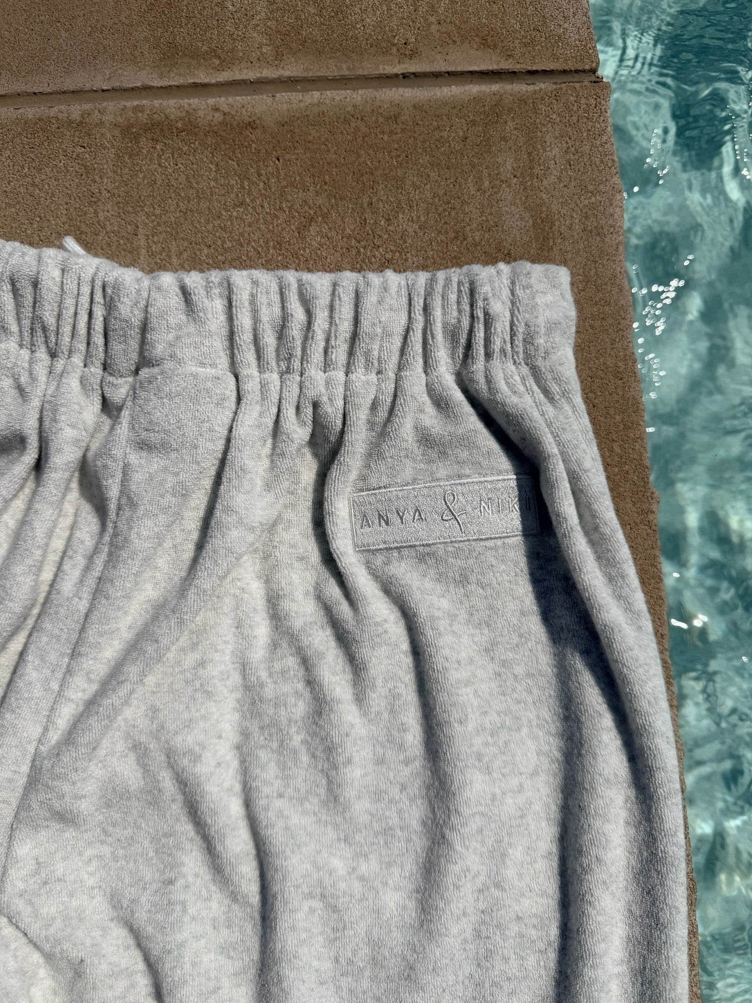Close-up of light gray heather sweatpants with an elastic waistband, laid next to a brown towel and a swimming pool.