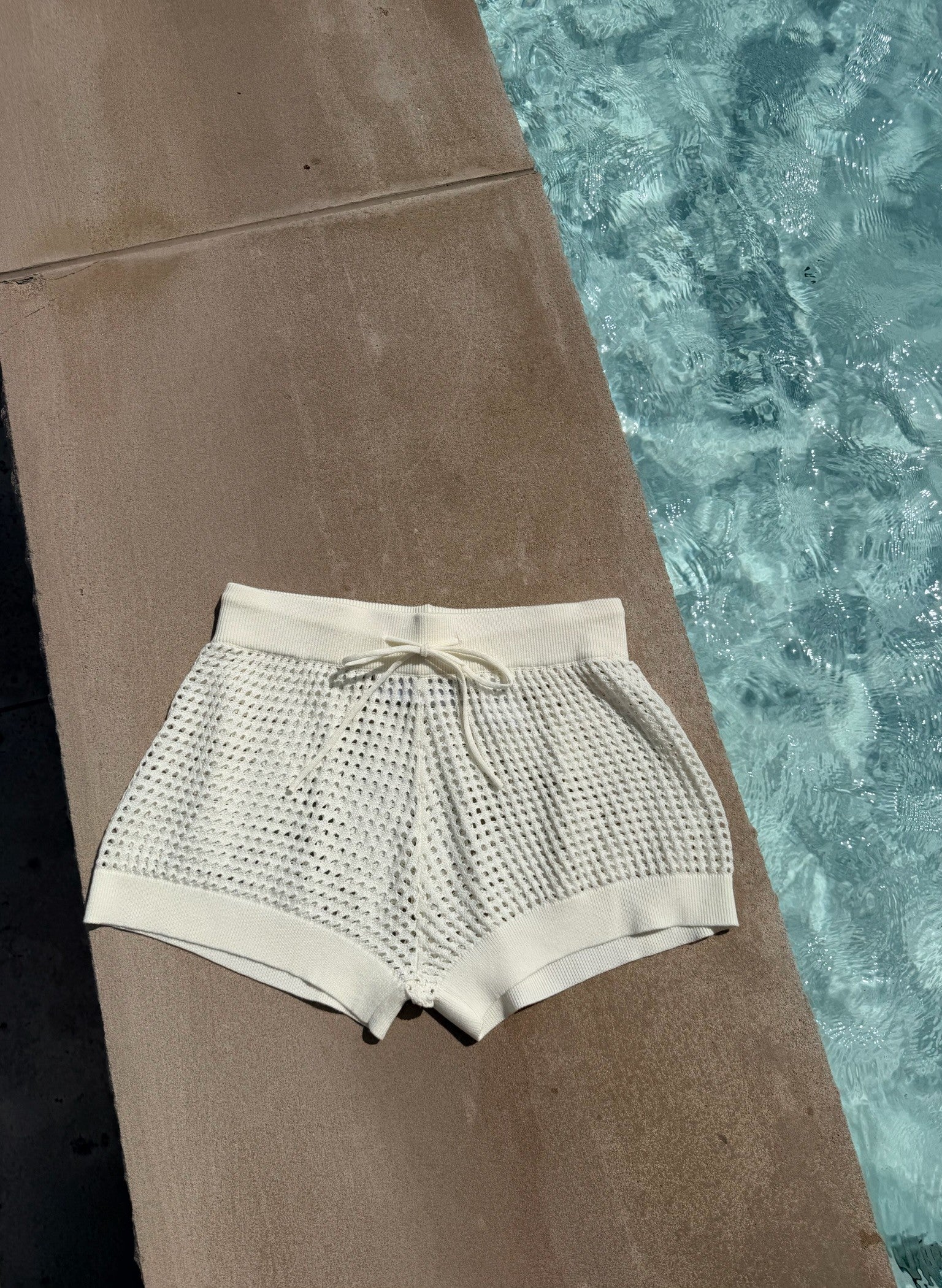 Cream crochet swim shorts with a drawstring waist, laid flat on a concrete pool deck next to a swimming pool.