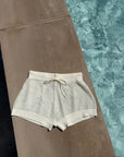 Cream crochet swim shorts with a drawstring waist, laid flat on a concrete pool deck next to a swimming pool.