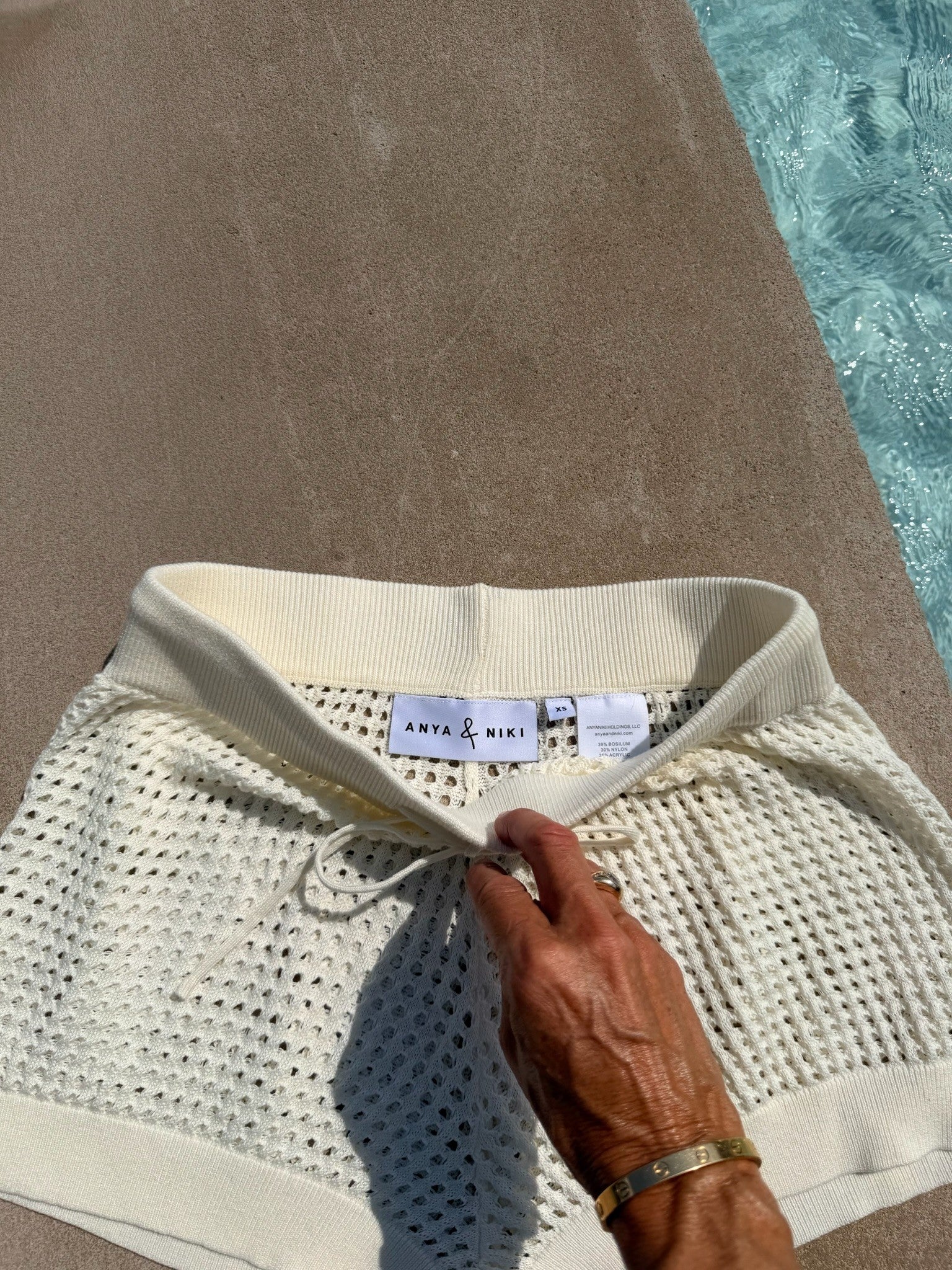 A close-up of a cream-colored knit swim cover-up with a wide ribbed waistband and drawstring, held by a hand. The brand label reads 'ANYA & NIKI'.
