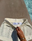 A close-up of a cream-colored knit swim cover-up with a wide ribbed waistband and drawstring, held by a hand. The brand label reads 'ANYA & NIKI'.