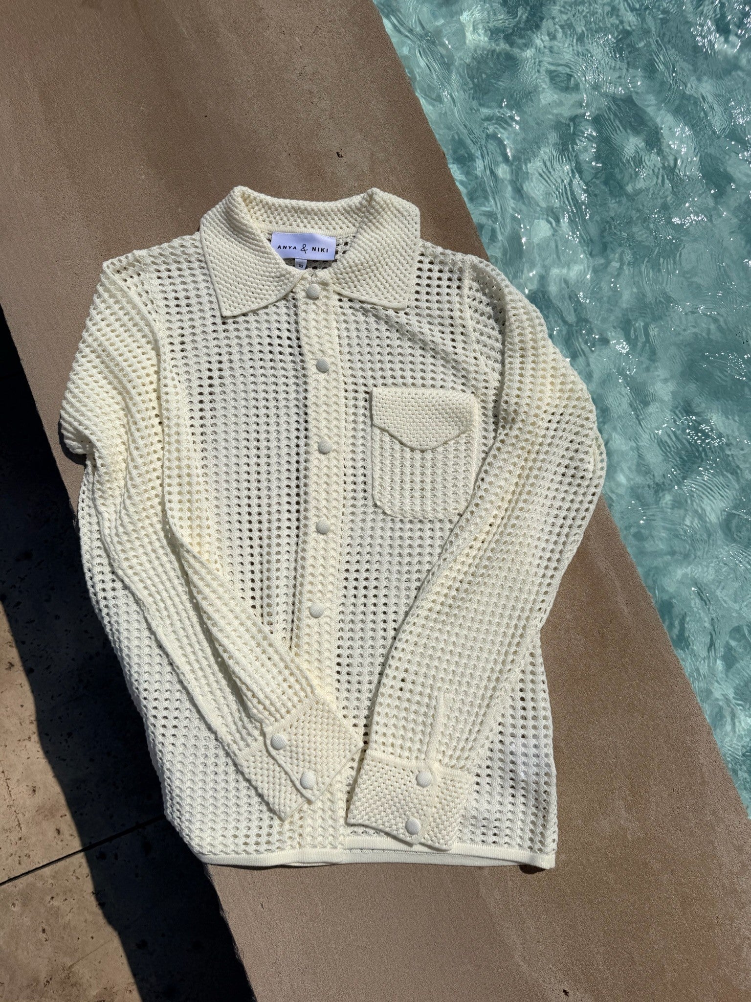 A cream-colored, open-knit long-sleeve button-up shirt with a collar and chest pocket, laid flat on a concrete surface next to a swimming pool.