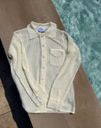 A cream-colored, open-knit long-sleeve button-up shirt with a collar and chest pocket, laid flat on a concrete surface next to a swimming pool.