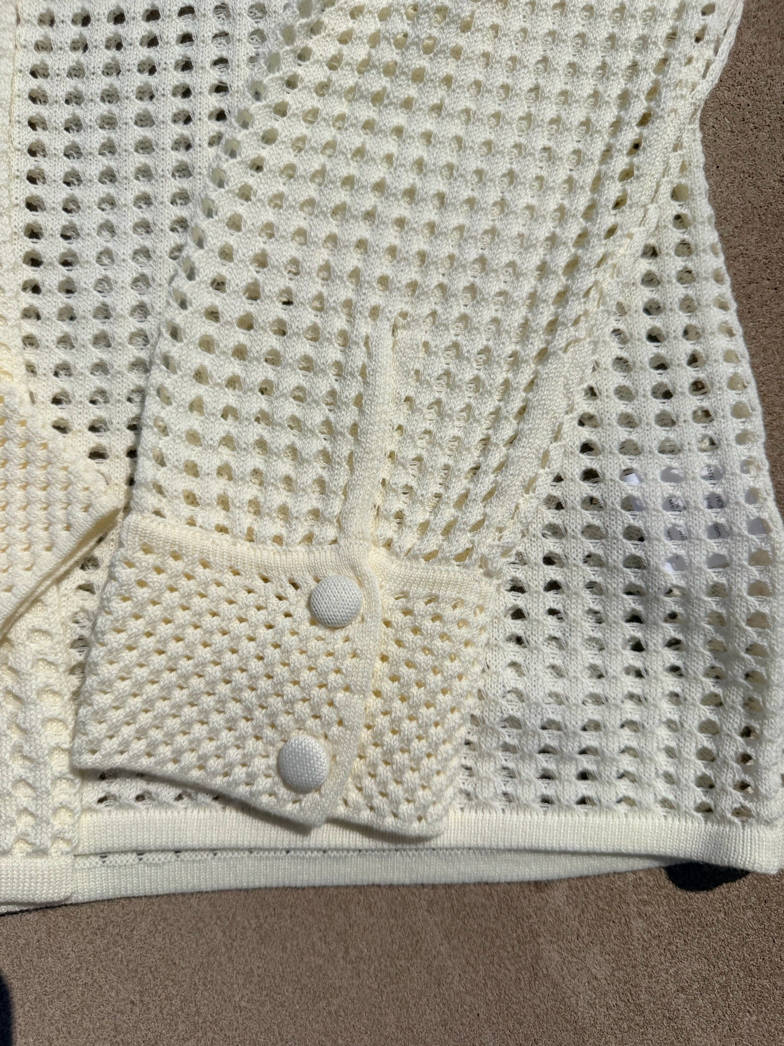 Close-up of a cream-colored open-knit cardigan or sweater with a buttoned cuff detail, laid flat on a brown surface.