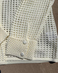Close-up of a cream-colored open-knit cardigan or sweater with a buttoned cuff detail, laid flat on a brown surface.