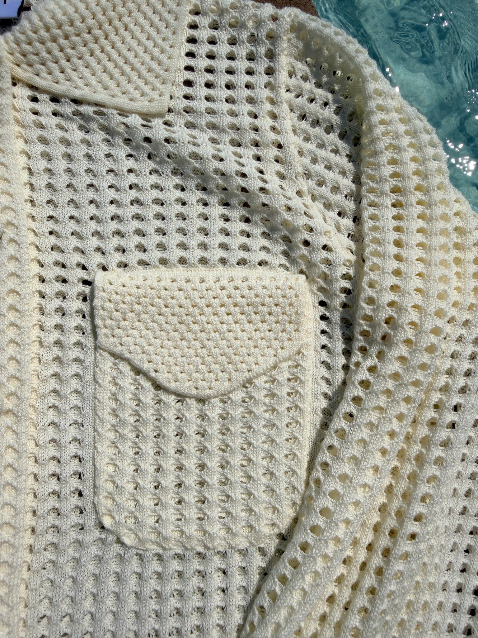 Close-up of a cream-colored open-knit cardigan sweater with a small chest pocket, showing the textured knit pattern and collar.