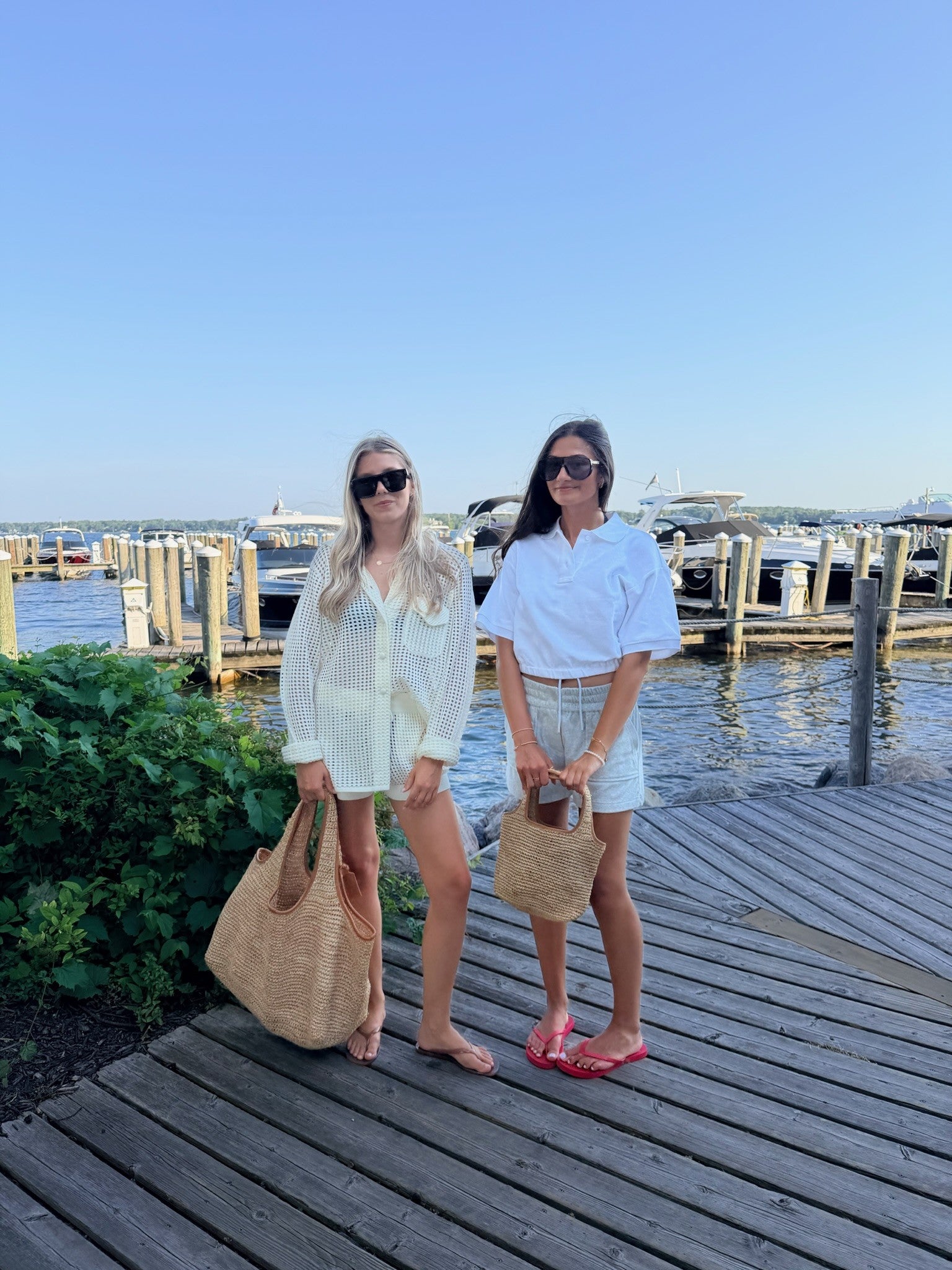 Two women standing on a wooden dock by the water, wearing sunglasses, light-colored outfits, and carrying large woven tote bags. Boats and a marina are visible in the background under a clear blue sky.