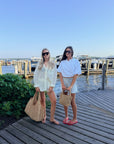 Two women standing on a wooden dock by the water, wearing sunglasses, light-colored outfits, and carrying large woven tote bags. Boats and a marina are visible in the background under a clear blue sky.