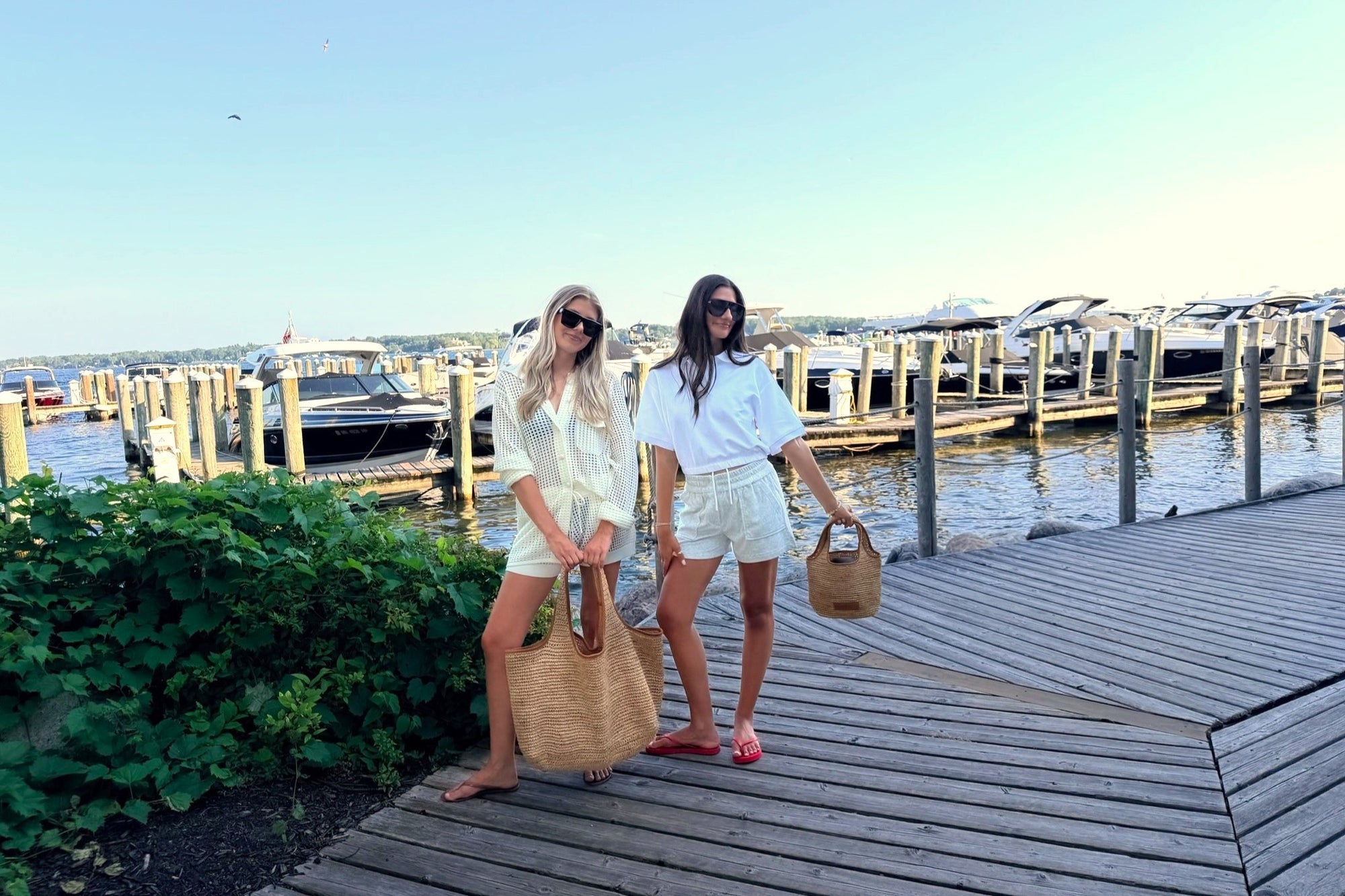 Two women standing on a wooden dock next to a marina with boats in the background. The woman on the left wears a cream crochet button-down shirt and shorts with a large beige tote bag, while the woman on the right wears a white polo shirt and shorts with a smaller beige tote bag.
