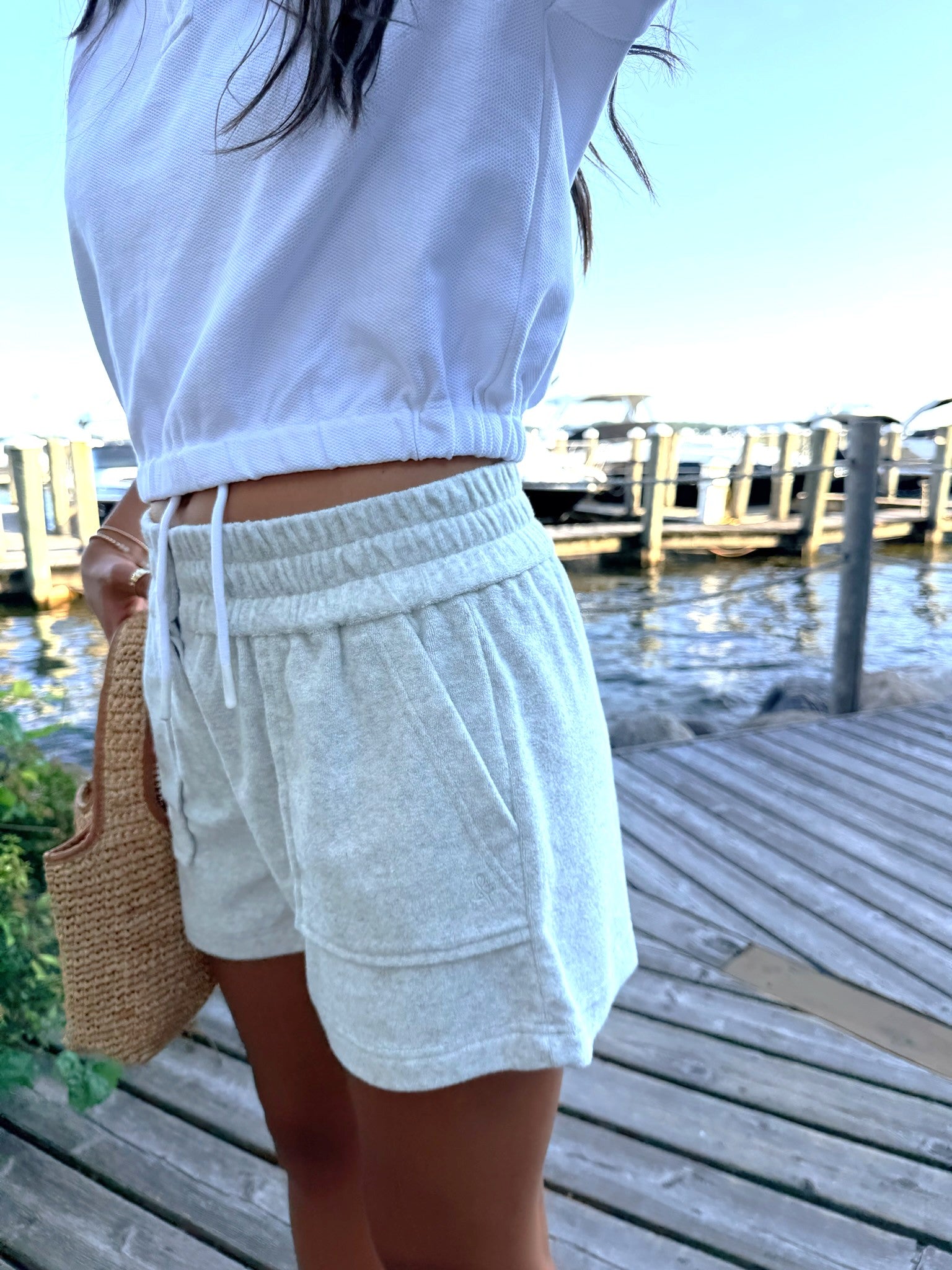 A person standing on a wooden dock by the water wearing light heather gray drawstring shorts and a white cropped top, holding a woven straw bag.