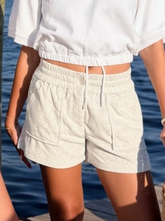 A person wearing light beige textured drawstring shorts and a white cropped top, standing outdoors with water in the background.