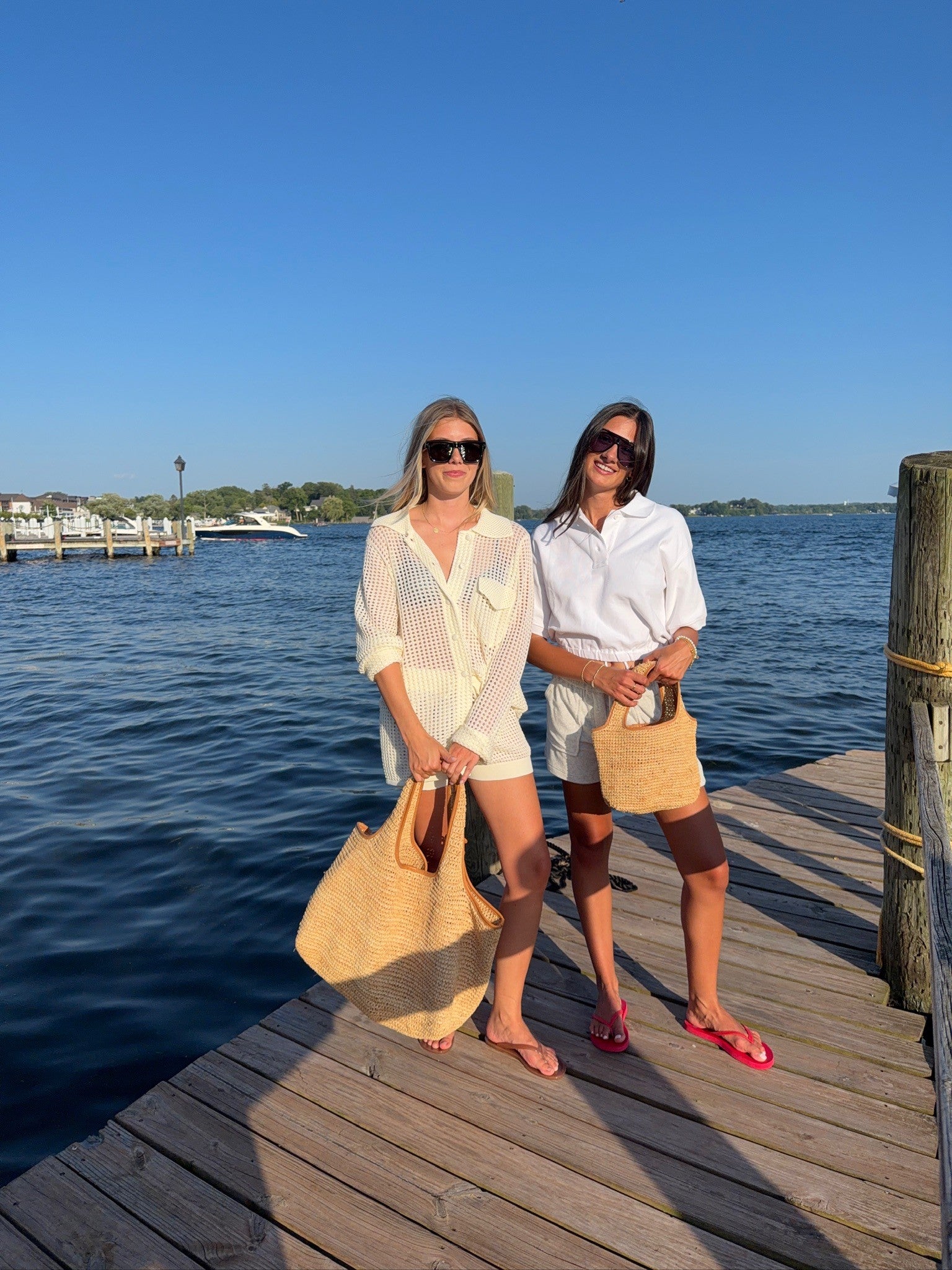 Two women standing on a wooden dock by the water, wearing sunglasses, white tops, and straw tote bags. The background features a clear blue sky and calm water.