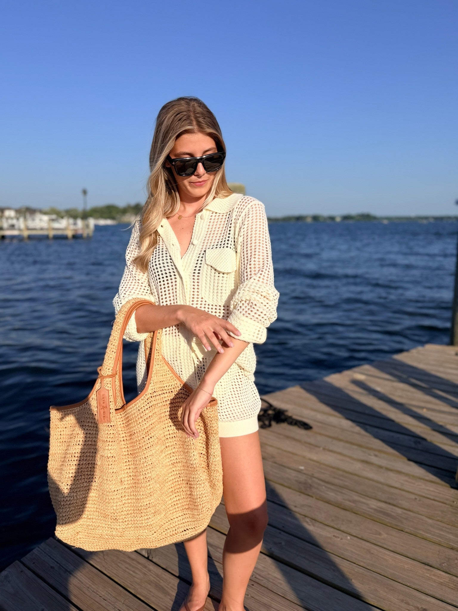 A woman stands on a wooden dock by the water wearing a cream-colored crochet knit shirt and shorts set, black sunglasses, and carrying a large woven straw tote bag.