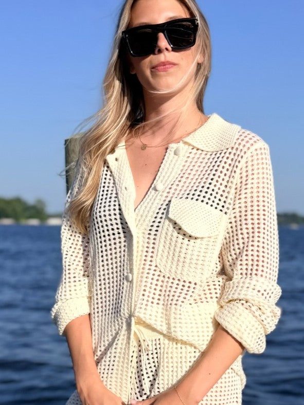 A woman wearing a cream-colored open-knit button-up shirt with a collar and chest pocket, styled with black sunglasses, standing outdoors with water and sky in the background.