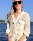 A woman wearing a cream-colored open-knit button-up shirt with a collar and chest pocket, styled with black sunglasses, standing outdoors with water and sky in the background.