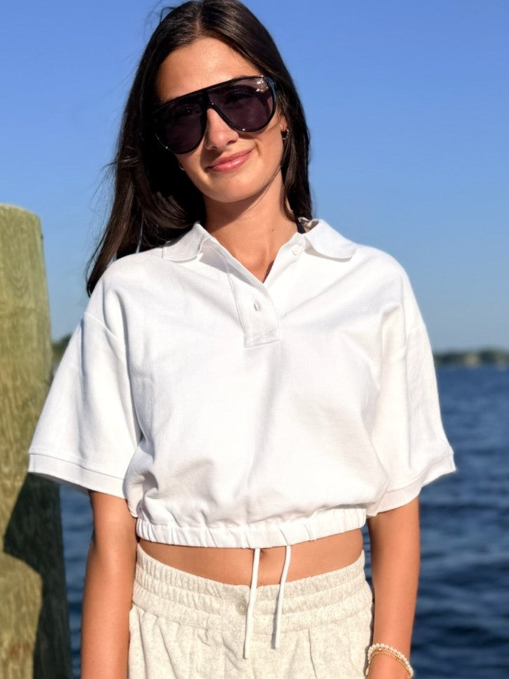 A woman wearing a white short-sleeve cropped polo shirt and light beige drawstring sweatpants, standing outdoors near the water with a wooden post on the left and blue sky and water in the background.