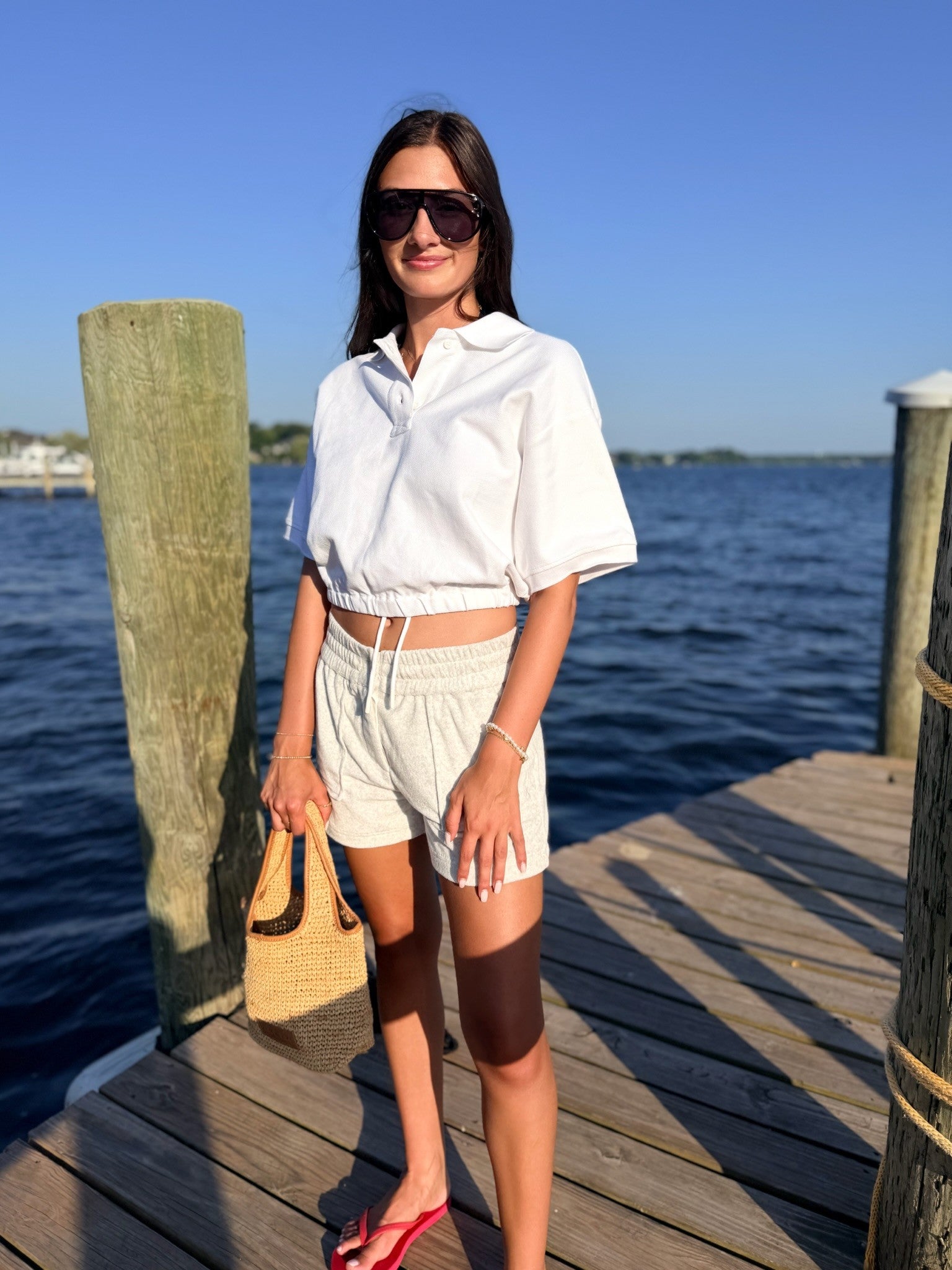 A woman stands on a wooden dock by the water wearing a white short-sleeve collared shirt, white shorts, sunglasses, and red sandals, holding a woven straw bag.