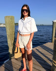 A woman stands on a wooden dock by the water wearing a white short-sleeve collared shirt, white shorts, sunglasses, and red sandals, holding a woven straw bag.