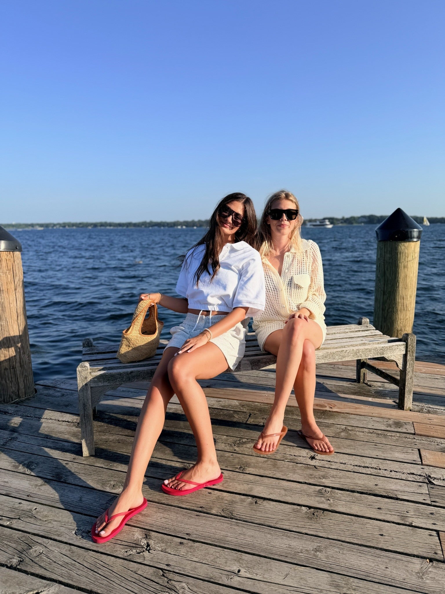 Two women sitting on a wooden dock bench with water and blue sky in the background. One wears a white shirt and shorts with red flip-flops and holds a tan sandal, while the other wears a cream lace-up top and shorts.